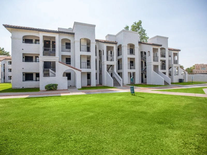 A white apartment complex with multiple units, featuring staircases leading to balconies. The surrounding area has green lawns and landscaped pathways.