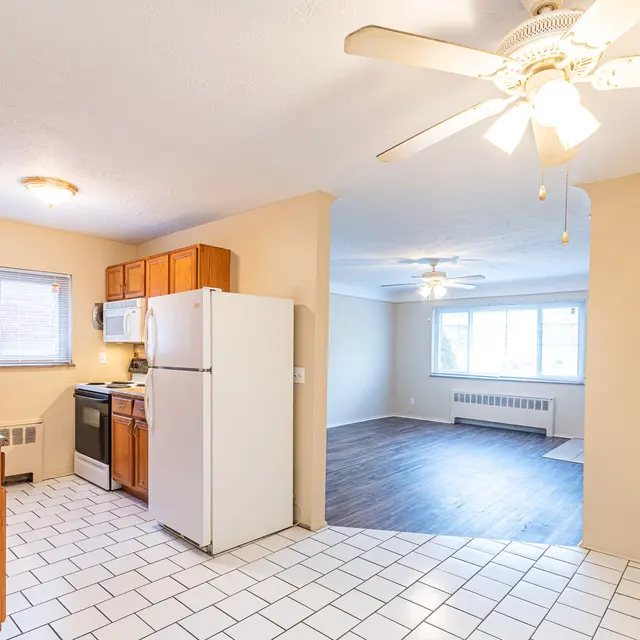 Spacious Kitchen and Living Room A spacious kitchen area leading to a living room. The kitchen has wooden cabinets, a white refrigerator, a stove, and white tiled flooring. The living room features large windows and a ceiling fan, with wooden flooring and neutral wall colors.