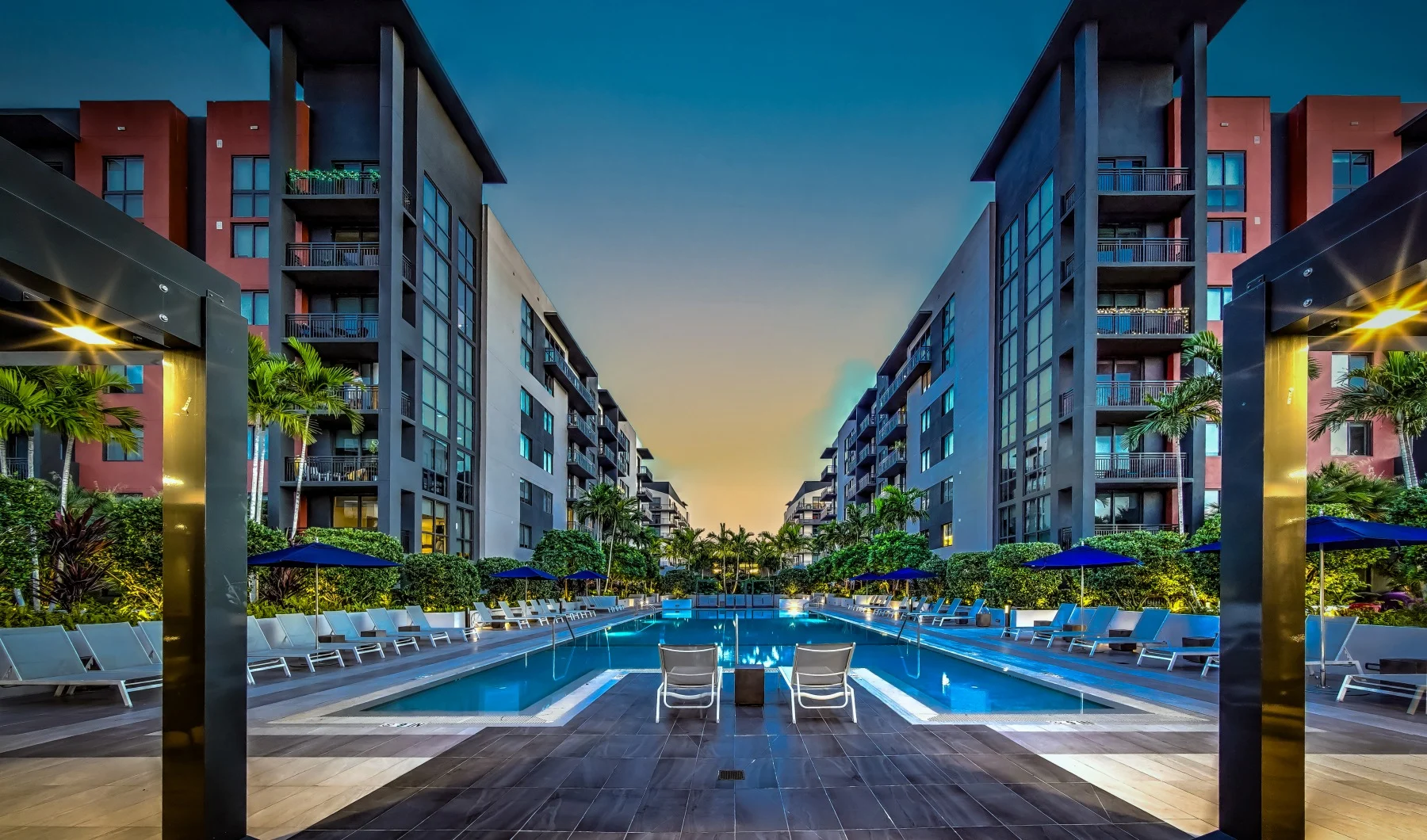 A modern apartment complex featuring a large swimming pool surrounded by lounge chairs and palm trees under a twilight sky.