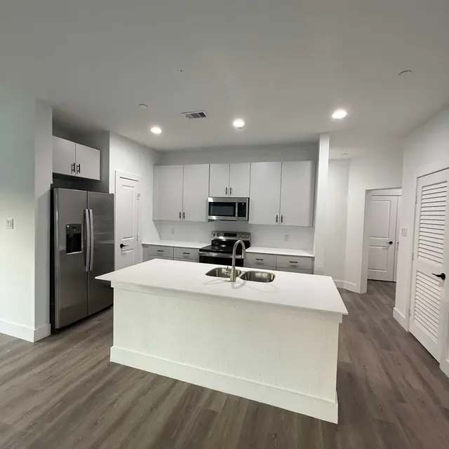 A modern kitchen featuring a large island, stainless steel appliances, and white cabinetry. The flooring is a light wood finish, and the walls are painted white, creating a bright and airy atmosphere.