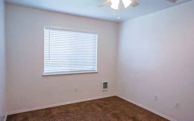 Empty Room with Natural Light A small, empty room with beige carpet and white walls. There is a window with blinds and a ceiling fan. The room is well-lit with natural light from the window.