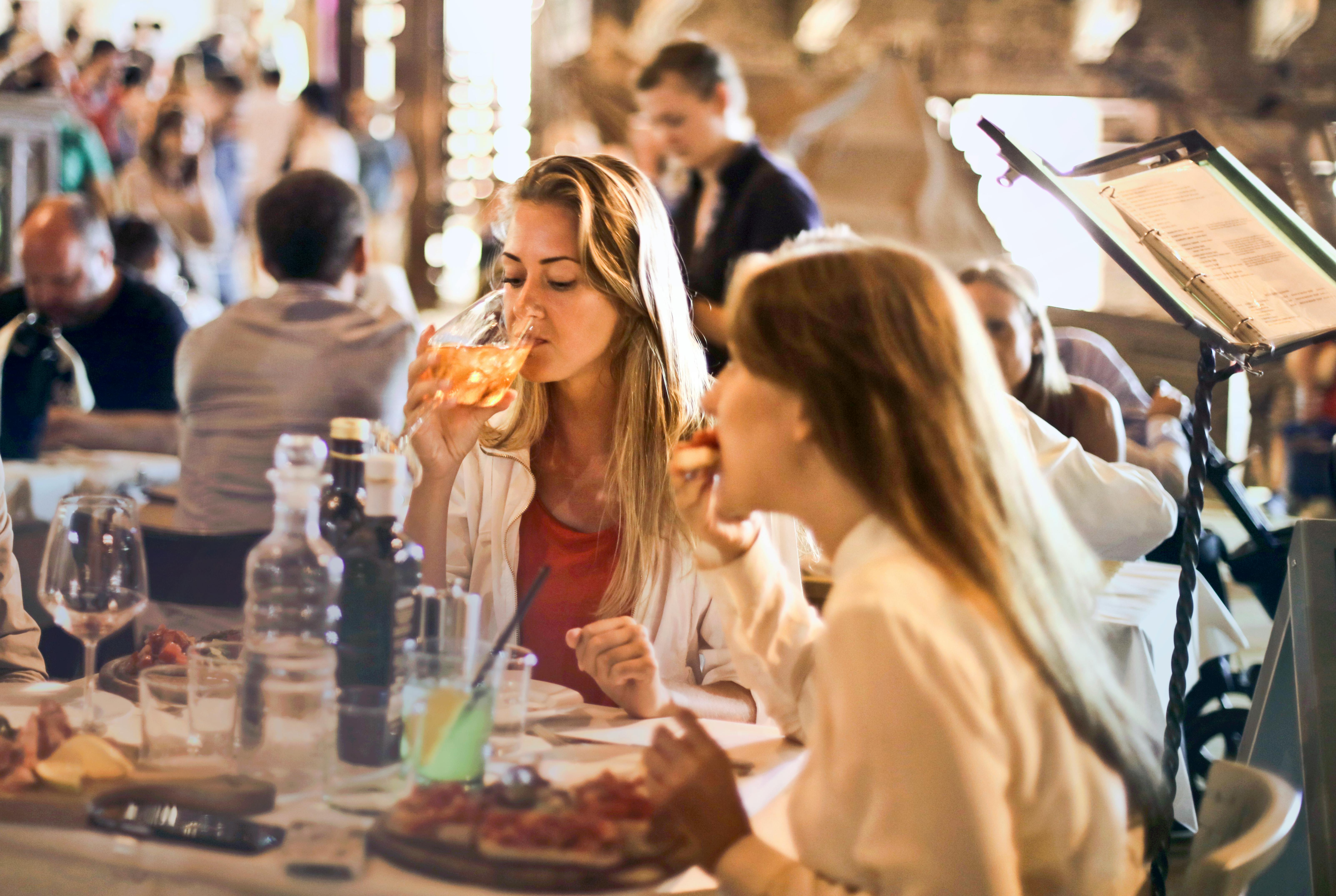 People dining in a bustling restaurant setting, with two women enjoying their drinks and food at a table.