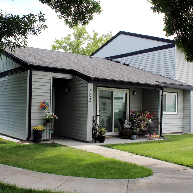 A modern single-story residence with a flat roof, featuring a landscaped front yard and porch. The house is painted in light gray with darker trim, and has a bike and potted plants on the porch. Soft green grass surrounds the home, with a sidewalk leading to the entrance.
