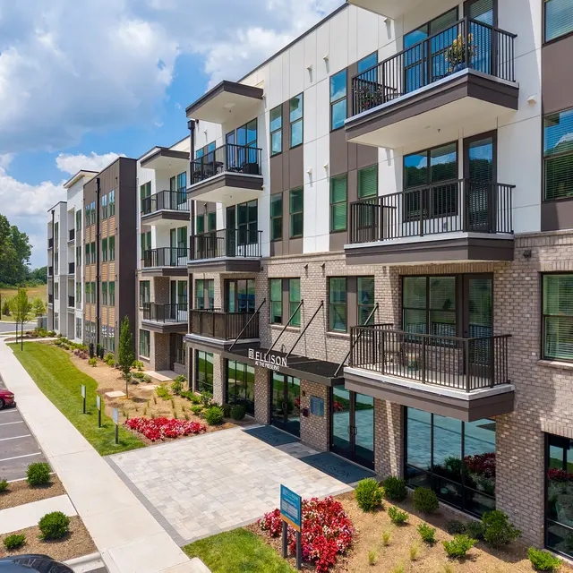A modern apartment complex featuring multiple stories with balconies and landscaped surroundings. The building has large windows and a clean, contemporary design. A parked car and open parking space are visible in the foreground.