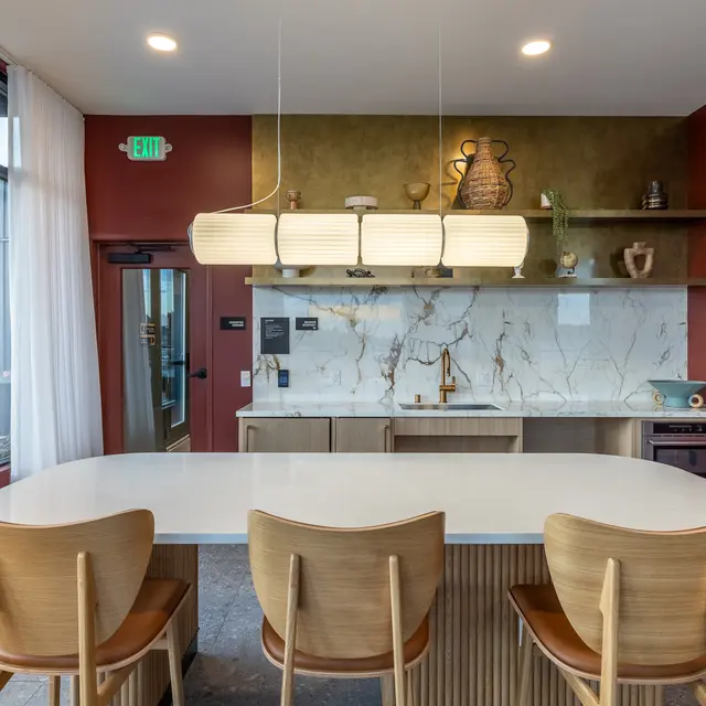 A modern kitchen interior featuring a large white countertop with wooden chairs, a stylish pendant light above, green textured walls, and a marble backsplash with decorative items.