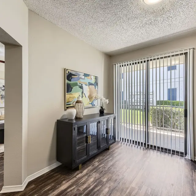 A view of a living space with an open layout leading to a sliding glass door, which opens to an outside area. The interior features light-colored walls, wooden flooring, and stylish decor including a cabinet and artwork. Soft lighting and a cozy atmosphere are evident.