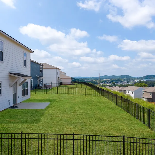 A spacious backyard with a well-maintained lawn, bordered by a black metal fence. Nearby houses are visible, and the landscape shows rolling hills under a bright blue sky with fluffy white clouds.