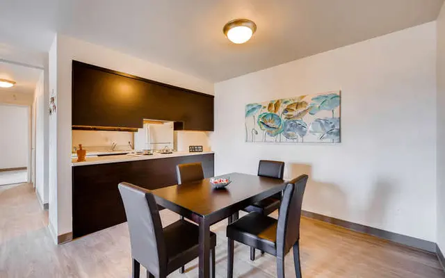 A modern dining area with a black table and chairs, adjacent to a kitchen with dark cabinetry, featuring wall art of blue and green flowers.
