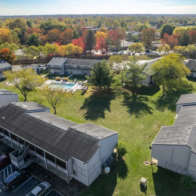Aerial view of an apartment complex with green lawns, trees, and a swimming pool, surrounded by colorful fall foliage.