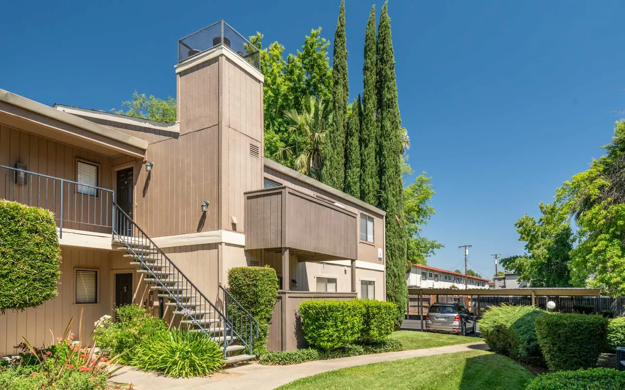 Exterior view of a multi-unit apartment building with a staircase, surrounded by greenery and blue sky.