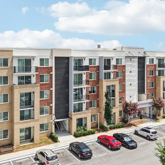 A modern apartment complex with multiple floors, featuring a mix of brick and siding exteriors. There is a spacious parking area in front with several parked cars and landscaped greenery visible around the building.