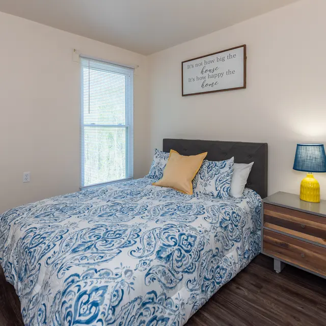 A cozy bedroom featuring a double bed with a blue and white patterned comforter, yellow accent pillow, and a bedside table with a blue lamp. A window allows natural light to enter, and wall decor is displayed above the bed.