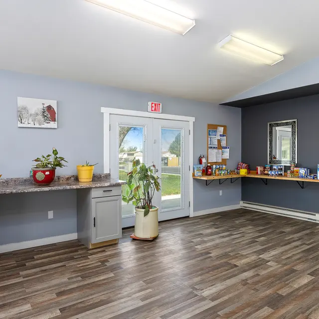 Interior Room with Kitchenette and Plants An empty room with wooden flooring, featuring a kitchenette with cabinets and various items on the counter. There are potted plants and a large set of doors leading outside. Walls are painted in light colors, with a few decorations.