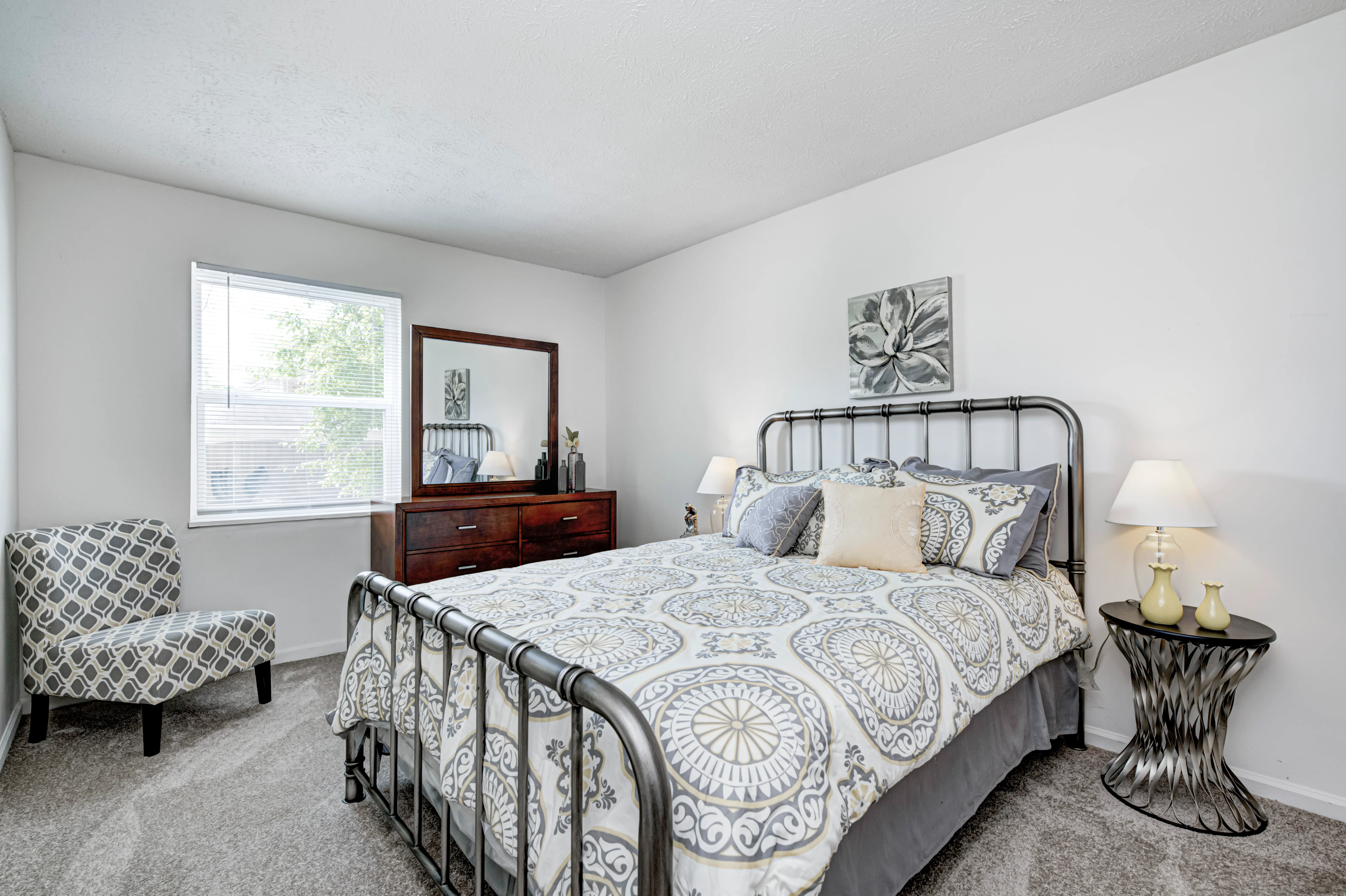 A modern bedroom featuring a queen-sized bed with a patterned bedspread, a wooden dresser, and a large mirror. The room has a chair in the corner, two bedside lamps, and a window providing natural light.