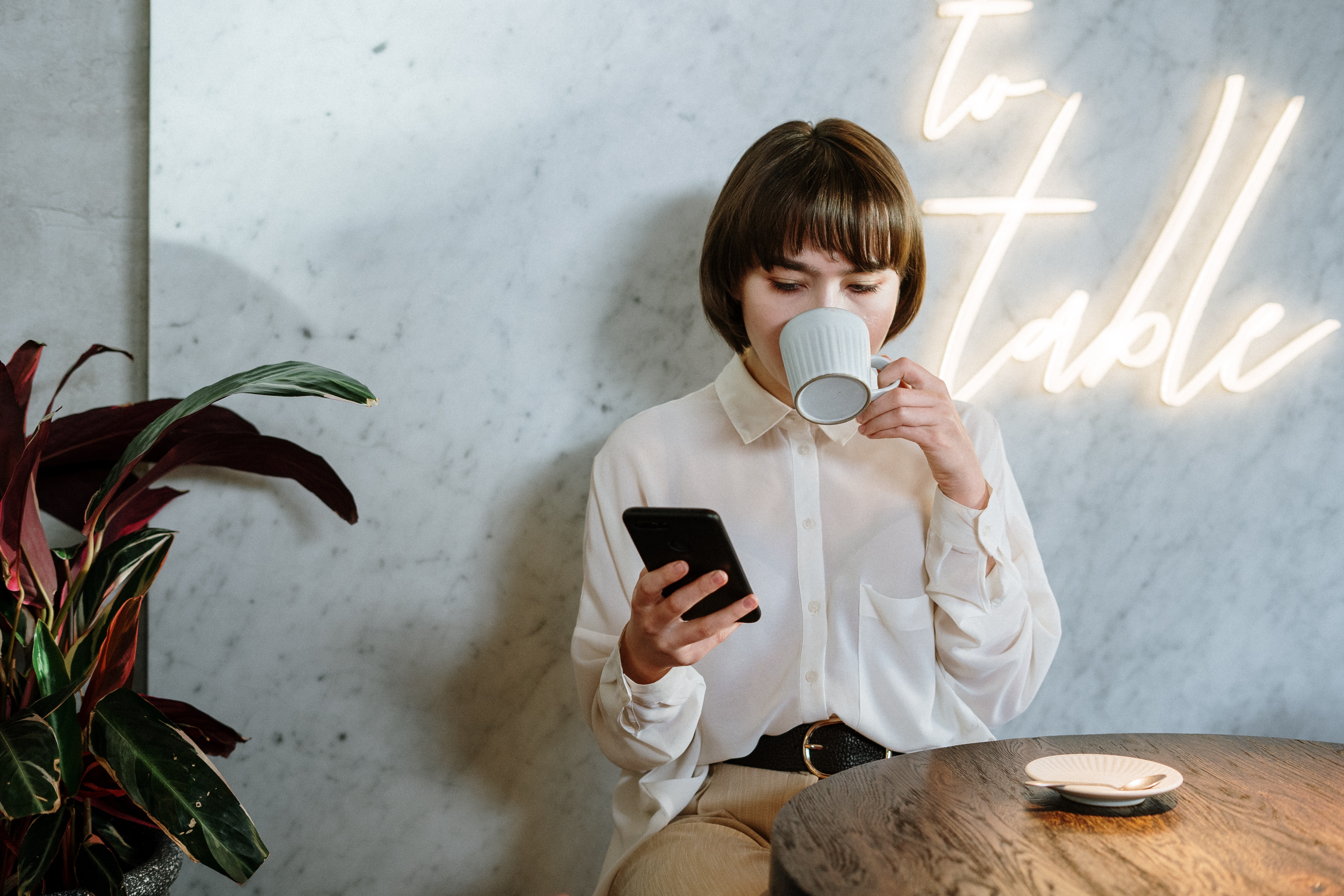Cafe Relaxation A woman sitting at a table in a cafe, holding a cup and looking at her phone. She wears a white blouse and is surrounded by a plant and a small plate on the table.