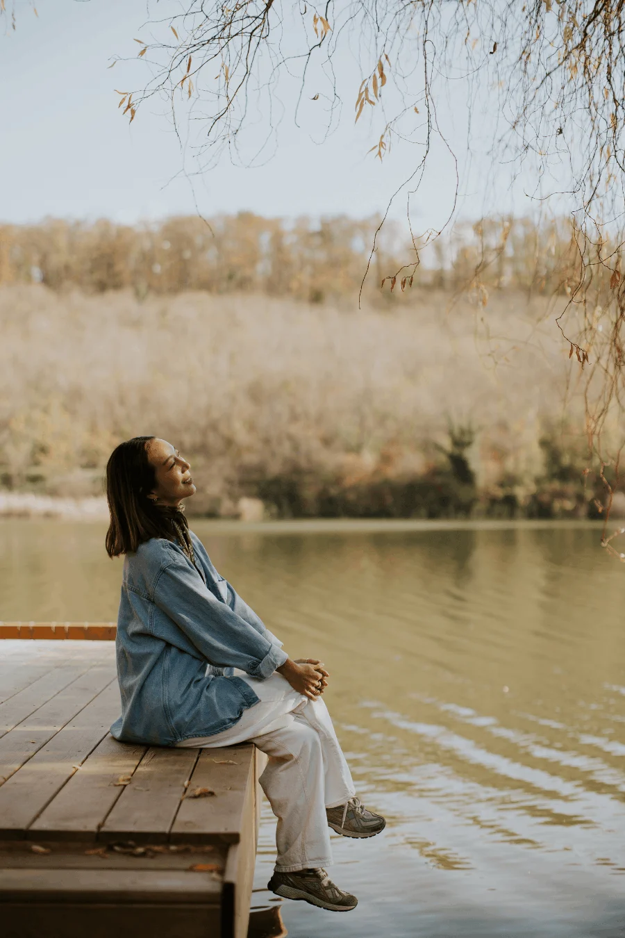 A person with long hair sitting on a wooden dock by a lake, gazing thoughtfully into the distance. They are wearing a blue jacket and white pants, surrounded by trees in autumn colors in the background.