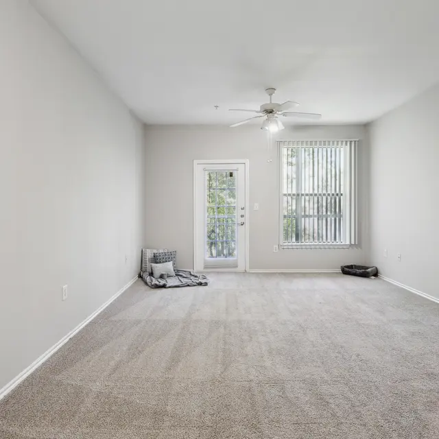 Empty Living Room Space A spacious empty living room with beige carpet, light gray walls, a ceiling fan, and a door leading to a small balcony with vertical blinds.