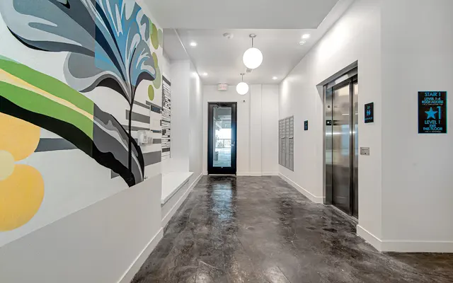 A modern hallway featuring an artistic wall mural, a glass door, elevator, and contemporary lighting fixtures.