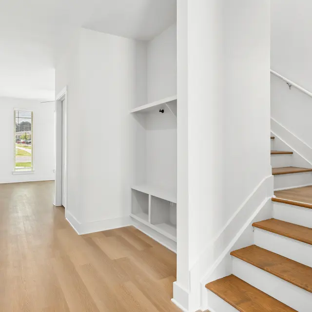 A spacious and bright entrance hallway featuring light wood flooring, white walls, and a staircase leading to the upper level. There is a built-in shelf on the right side and a window letting in natural light.