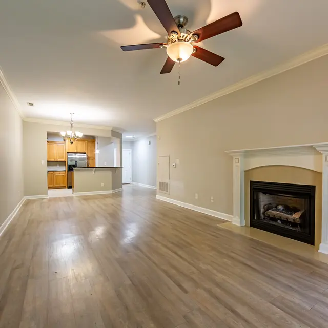 A spacious living room featuring hardwood flooring, a ceiling fan, and a fireplace. The room is painted in neutral tones, leading into a kitchen area visible in the background.
