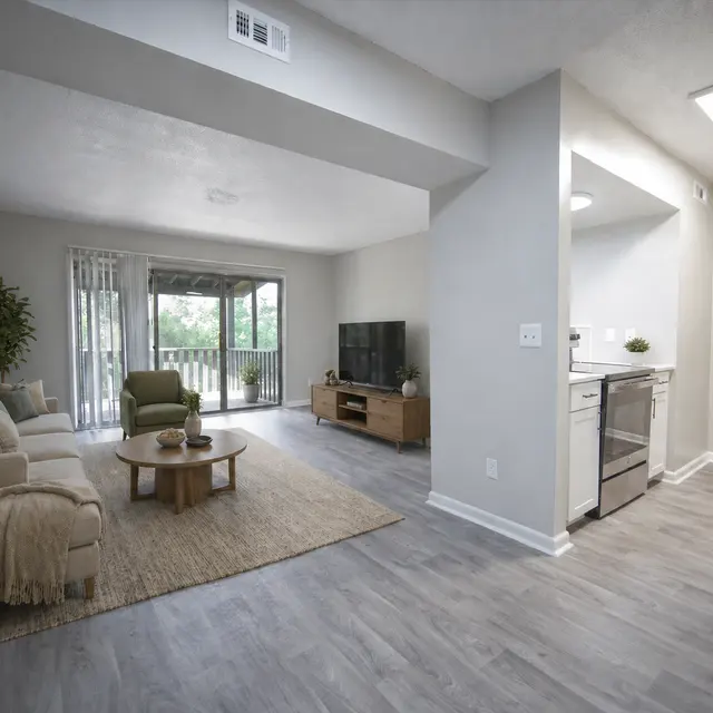 A bright and modern living area featuring a light-colored sofa, a small coffee table, a green accent chair, and a television. The space has large windows with curtains, allowing natural light to fill the room. To the right, there is an open kitchen with stainless steel appliances.