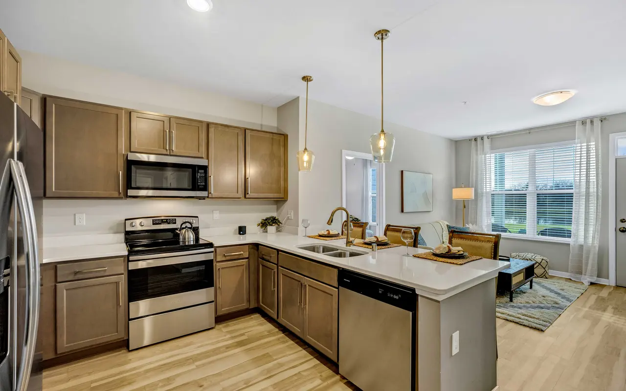 A modern kitchen featuring stainless steel appliances, wooden cabinets, and an island with a sink. The kitchen opens to a bright living area with a seating arrangement.