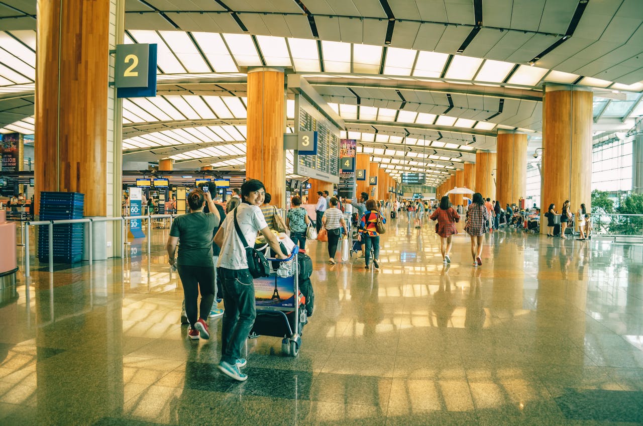 A busy airport terminal with travelers walking, some pushing luggage trolleys. Bright and spacious interior with tall pillars and overhead signs indicating gates.