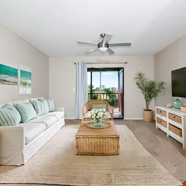 Bright and airy living room featuring a light-colored sofa with decorative pillows, a wicker coffee table, and a TV on a modern white cabinet. A sliding door opens to a balcony with a view. Indoor plants add a touch of greenery.