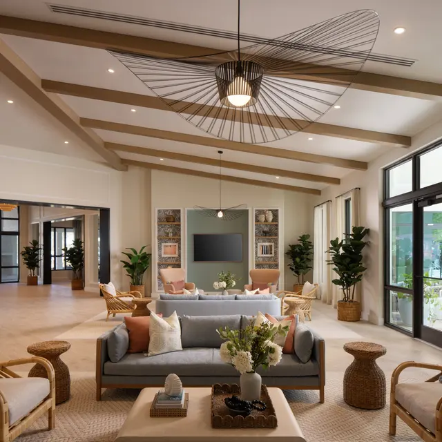 A spacious and elegantly designed living room featuring light wooden beams on the ceiling, contemporary furniture, and large windows letting in natural light. Decorative plants are placed around the room, enhancing the cozy atmosphere.