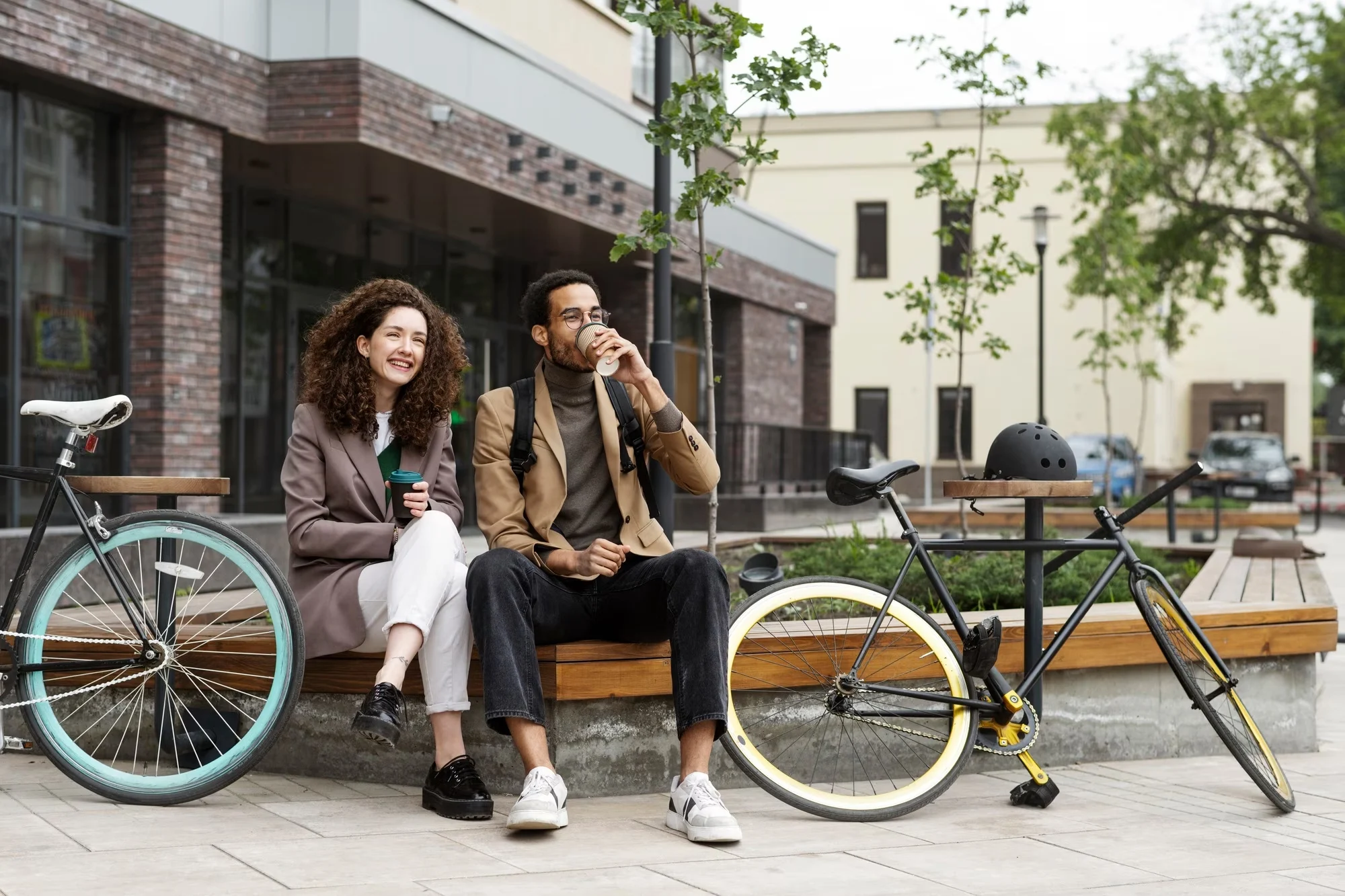 A young couple sitting on a bench with bicycles beside them, enjoying drinks in a modern urban setting.
