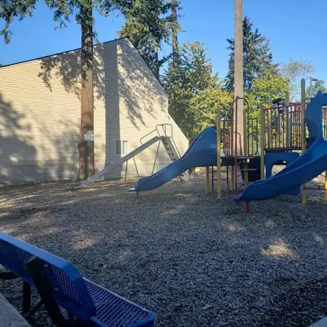 A playground area featuring a blue slide, a climbing structure, and a swing set, with benches nearby and an apartment building in the background under a clear sky.