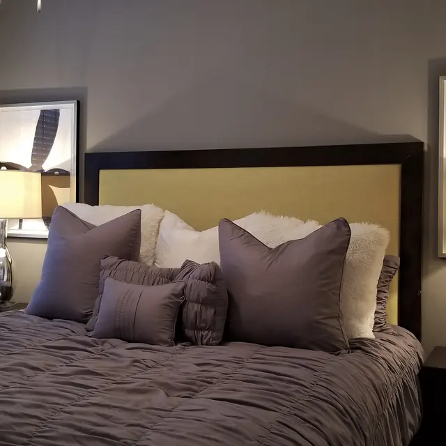 A cozy modern bedroom featuring a large bed with multiple pillows, a patterned comforter, and stylish bedside lamps. The walls are a soft color, and there are framed artworks on either side of the bed.