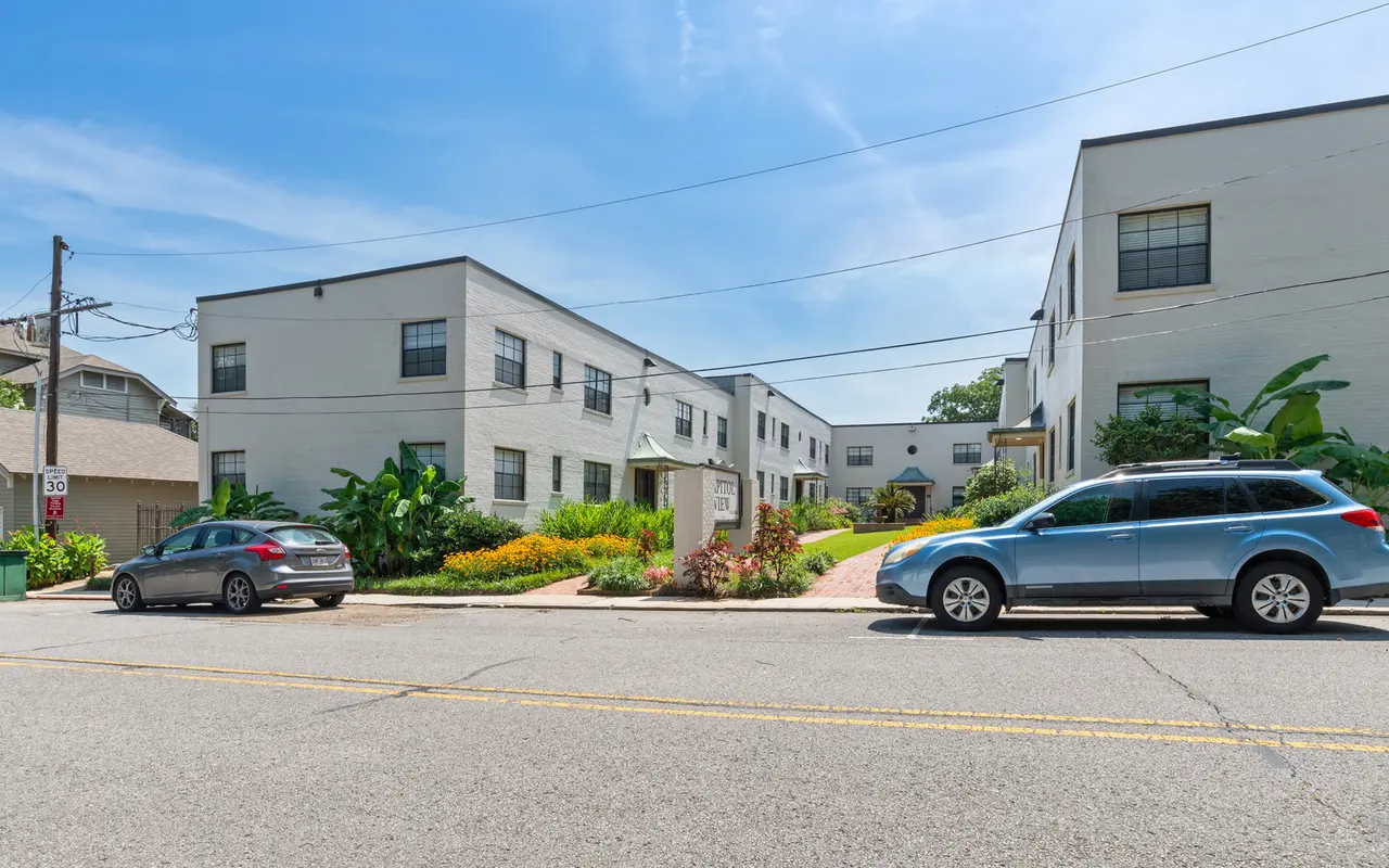 A view of a multi-unit apartment complex from the street with greenery and parked cars in front.