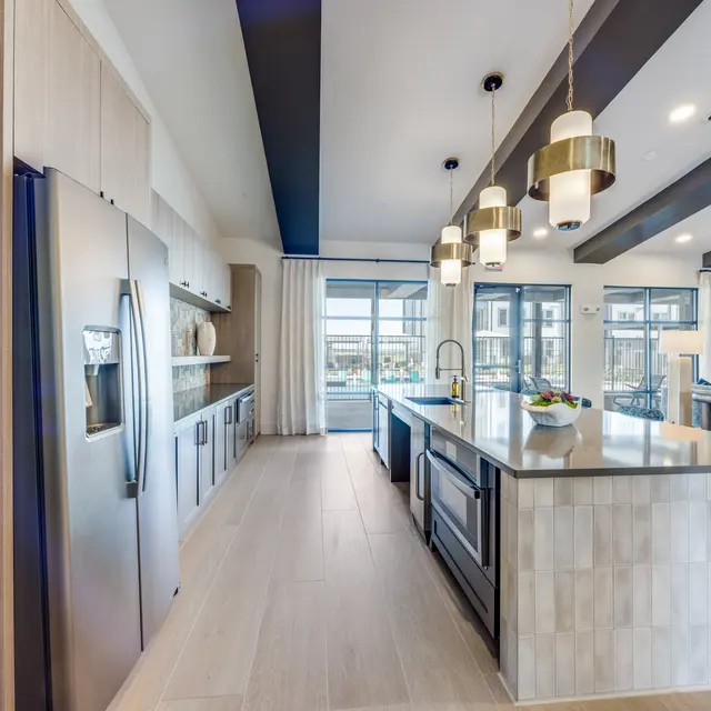 A spacious, modern kitchen featuring sleek stainless steel appliances, wooden cabinetry, and elegant pendant lighting. The island is adorned with a simple centerpiece, while large windows allow for abundant natural light and a view outside.