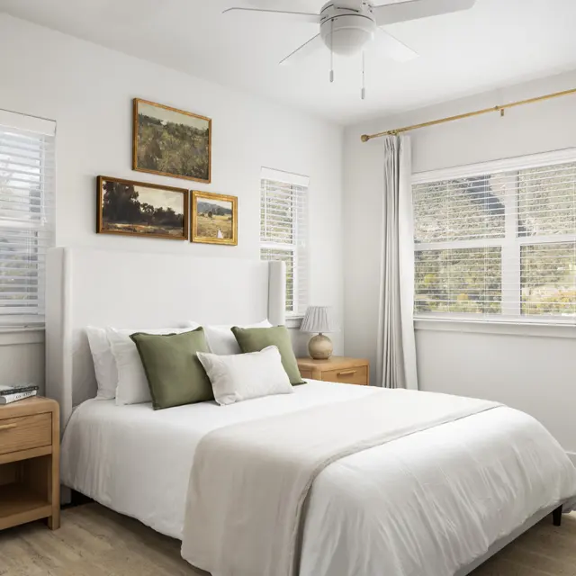 A modern and elegant bedroom featuring a queen-sized bed with white linens and green accent pillows. There are two nightstands with lamps and framed artwork on the wall. A large window is dressed with light curtains, letting in natural light.