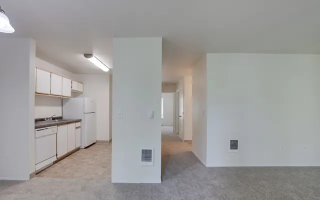 An interior view of a spacious, minimalist apartment featuring a kitchen area and a carpeted living space.