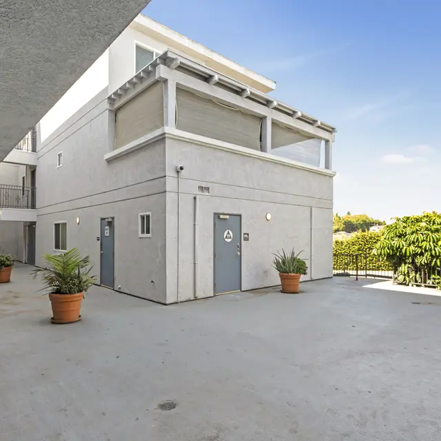 A spacious courtyard area of an apartment complex with a smooth concrete floor and potted plants. The building features light-colored walls and blue doors, with some greenery visible in the background.