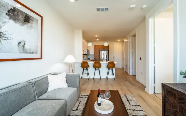 A modern living room featuring a grey sofa, a wooden coffee table with wine glasses, a bottle of wine, and a bowl of snacks. In the background, there are three bar stools and an open kitchen area with pendant lighting. A framed artwork is hanging on the wall.