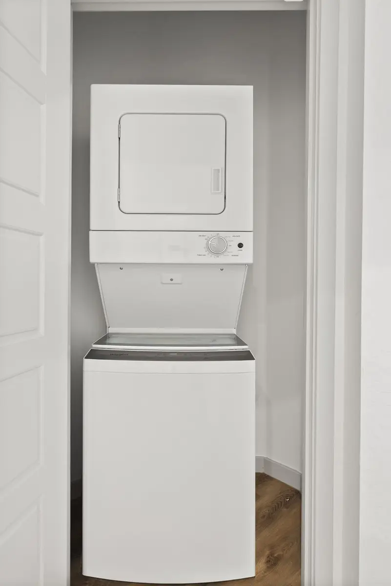 Stacked white washer and dryer in a narrow laundry space with light walls and wooden flooring.