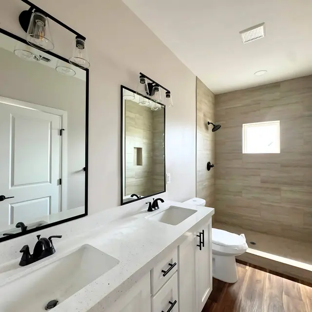 A bright and modern bathroom featuring dual sinks with a white countertop, two large mirrors, a beige tiled shower area, and dark fixtures.