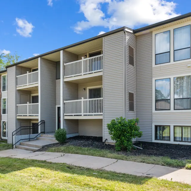 A modern three-story apartment building with balconies, surrounded by green grass and trees under a blue sky with white clouds.