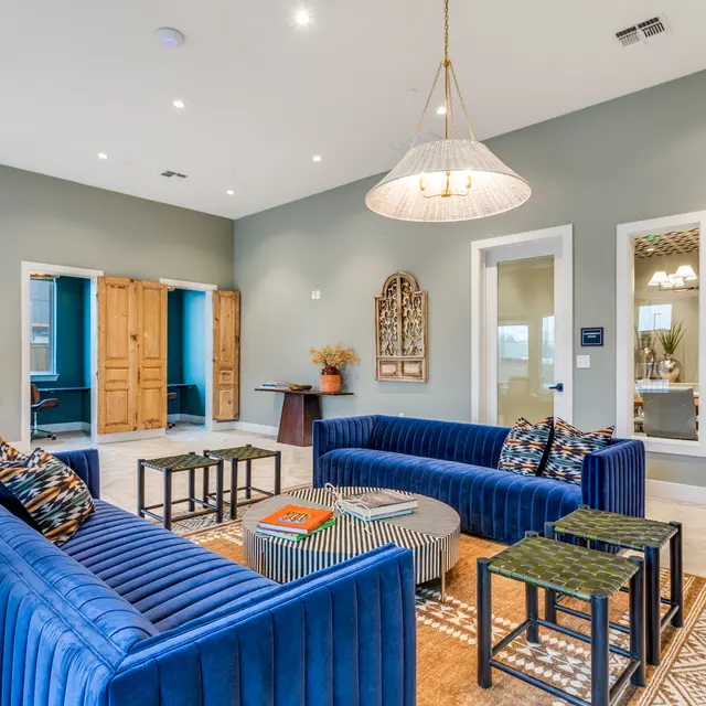 A modern lounge area featuring blue sofas, a patterned rug, coffee tables, and a chandelier. Brightly lit with large windows and stylish decor.