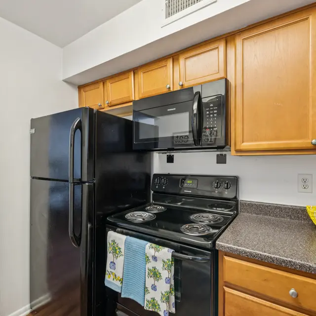 Cozy Modern Kitchen A modern kitchen featuring wooden cabinets, a black refrigerator, a black microwave, and a black stove. Countertops are gray with a decorative yellow bowl and kitchen utensils. There is a towel hanging on the stove.