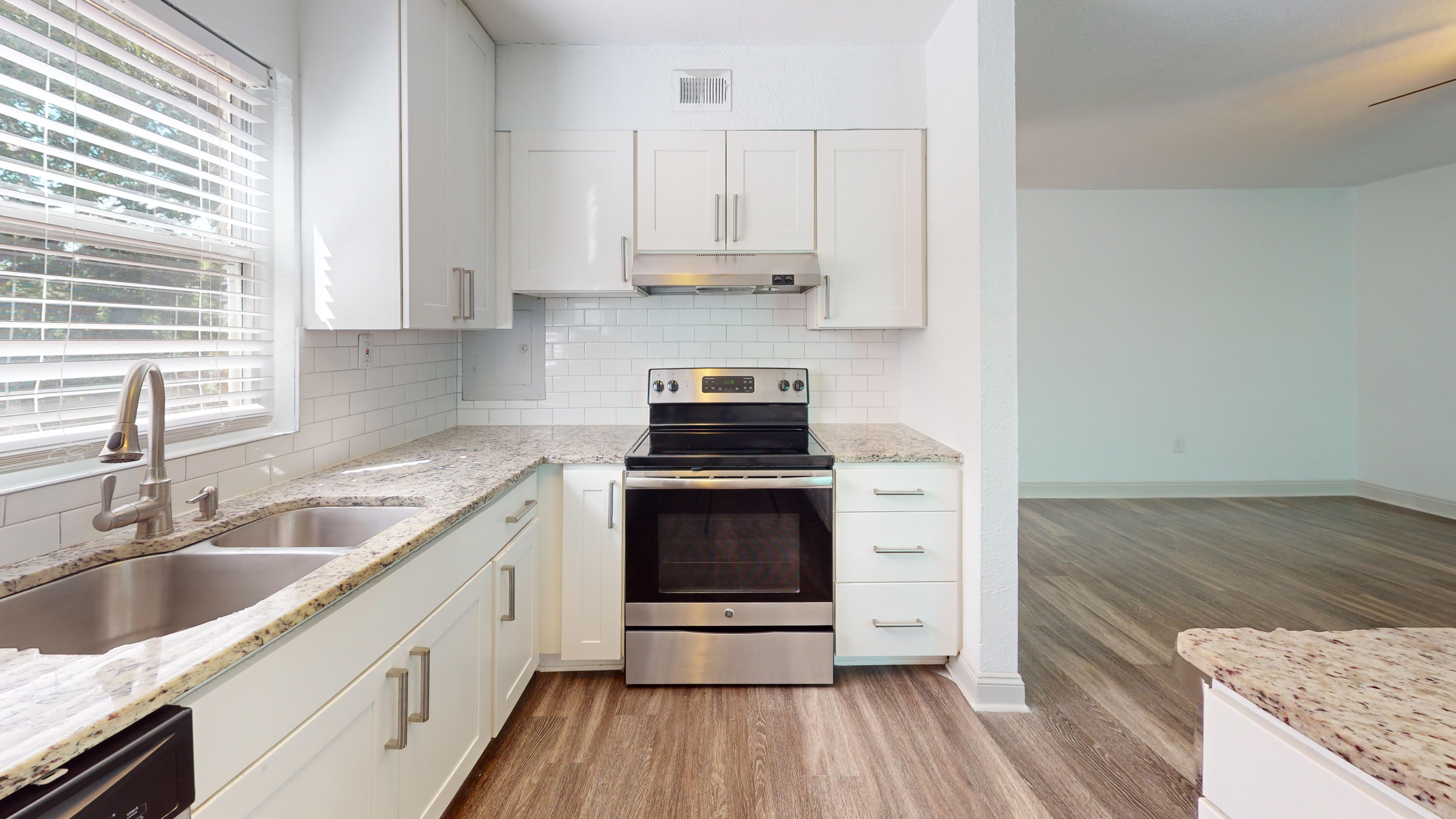 Stylish Modern Kitchen A modern kitchen with white cabinets, stainless steel appliances, and a spacious countertop. Natural light streams in through the window, highlighting the wooden floor and tile backsplash.