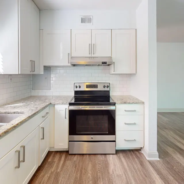 A modern kitchen with white cabinets, stainless steel appliances, and a spacious countertop. Natural light streams in through the window, highlighting the wooden floor and tile backsplash.