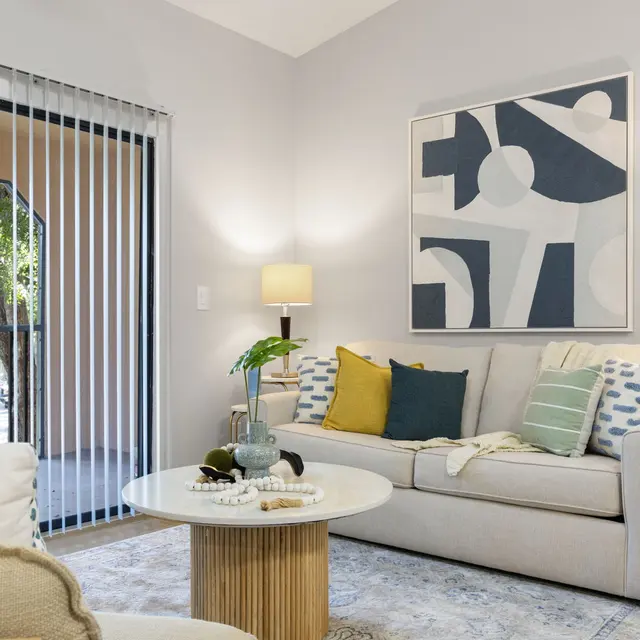 A modern living room featuring a light-colored sofa adorned with colorful cushions. There's a round coffee table with decor items in the center, and a textured rug on the floor. The walls are painted in a soft gray, complemented by a large abstract painting. A lamp provides warm lighting, and a sliding glass door reveals natural light from outside.