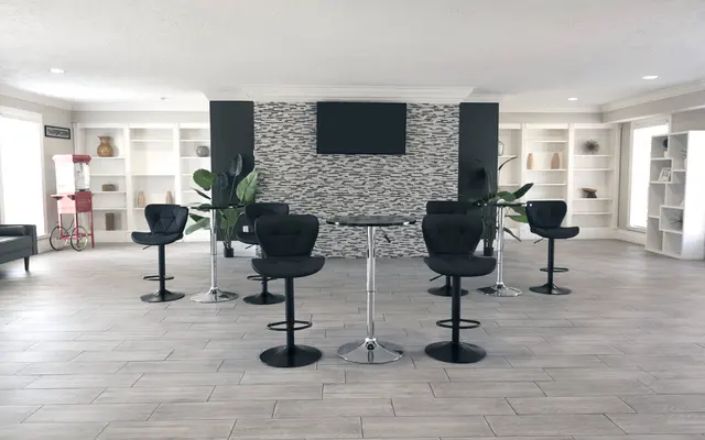 A spacious modern lobby area featuring six black barstools arranged around a sleek circular table, with a stone wall backdrop and decorative shelves filled with various items. There are two indoor plants and a popcorn machine visible in the background.