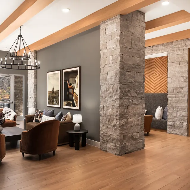 A modern living room featuring a cozy seating area with brown armchairs, a coffee table, and decorative artwork on the walls. Large windows allow natural light to shine in, while a stylish chandelier hangs from the ceiling. A stone column adds texture to the room, and there is a nook with built-in shelving visible in the background.