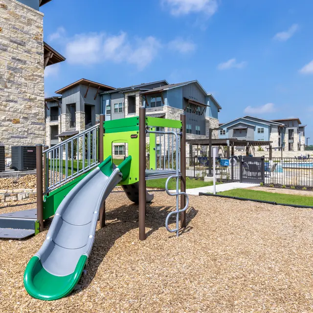 A playground featuring a gray and green slide, climbing structure, and surrounded by gravel. In the background, there are modern residential buildings and a fenced area visible under a clear blue sky.