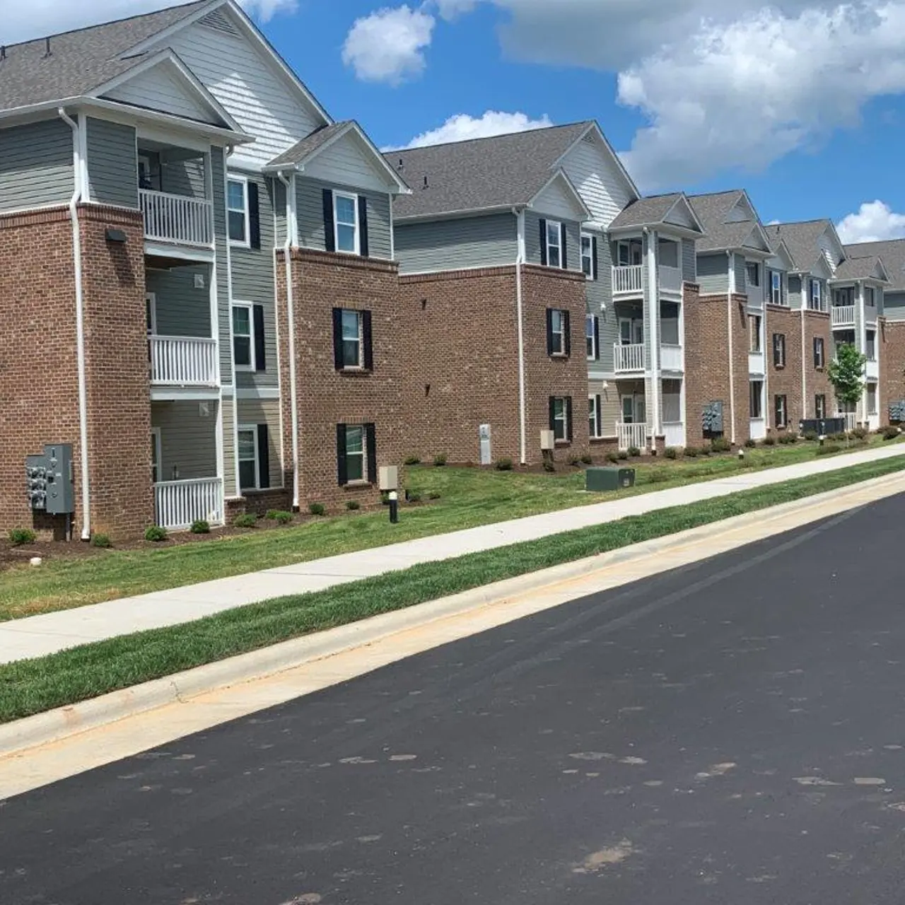 A row of multi-story apartment buildings with brick and siding exteriors. The foreground shows a paved road lined with grass, trees, and sidewalks.